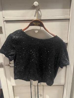 Svart paljettopp med snörning - Snygg croppad svart topp täckt med glittriga paljetter. Toppen har korta ärmar, rund halsringning och öppen rygg med snörning baktill. Perfekt för dig som vill sticka ut och glänsa lite extra.