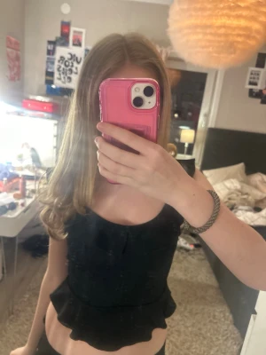 Svart volang topp med breda axelband - Säljer en svart croppad topp med breda axelband och volangkant nedtill. Toppen har en figurnära passform och är perfekt för dig som gillar en trendig och enkel stil. Materialet känns mjukt och stretchigt, vilket gör den bekväm att bära.