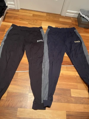 Två par mjukisbyxor från BOSS - Säljer två par mjukisbyxor från BOSS, ena i svart och den andra i mörkblått, båda med grå sidopaneler och resår i midjan samt benslut. Perfekta för chill dagar eller när du vill ha det bekvämt. Diskret BOSS-logga på vänster sida.