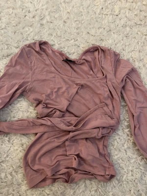 Rosa omlottopp från Gina Tricot - Snygg rosa omlottopp från Gina Tricot i mjuk bomull. Toppen har långa ärmar och ett knytband i midjan som ger en feminin siluett. Perfekt att styla med jeans eller kjol för en trendig look.