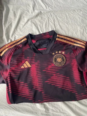 Äkta (original) Tysklands matchtröja Adidas svart/röd - Säljer en officiell Tyskland matchtröja från Adidas i svart och rött med guldiga detaljer. Tröjan har Tysklands landslagsemblem och fyra stjärnor på bröstet, samt Adidas-logga och klassiska ränder på axlarna. Tillverkad i lätt och ventilerande material.