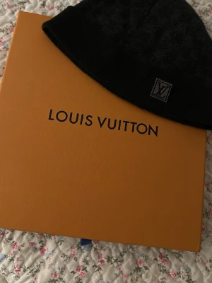 Svart Louis Vuitton mössa - Snygg svart mössa från Louis Vuitton med diskret rutmönster och vikt kant. Märkets klassiska LV-logga sitter framtill på en patch. Tillverkad i mjukt och varmt material som passar perfekt för kyliga dagar. Med tillhörande låda