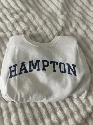 Vit sweatshirt från 157 med tryck - Säljer en vit sweatshirt från lager 157 i storlek XS med mörkblått HAMPTON-tryck på bröstet. Tröjan har rund halsringning och är gjord i mjukt, fleeceliknande material på insidan. Perfekt för dig som gillar college-stil och enkel design.