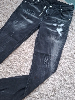 Svarta Dsquared2 jeans - Säljer ett par svarta skinny jeans med slitningar och hål på låret och knät. Jeansen har en cool dragkedjedetalj vid fickan och en tvättad look med vita partier. Perfekta för dig som gillar streetstyle och en edgy vibe.