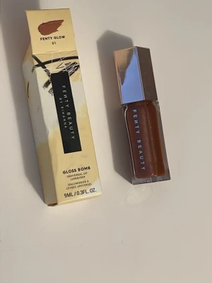 Fenty Beauty Gloss Bomb Fenty Glow - Fenty Beauty Gloss Bomb i färgen Fenty Glow är ett läppglans med en varm, nude-brun ton och subtilt skimmer. Kommer i en stilren, sexkantig plastförpackning med spegelblankt lock. Innehåller 9 ml och ger en glansig finish som känns lyxig och trendig.