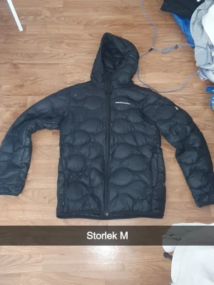 Svart dunjacka med huva från Peak Performance - Säljer en svart dunjacka från Peak Performance med huva och dragkedja framtill. Jackan har en quiltad design och diskret logga på bröstet. Perfekt för kalla dagar och riktigt skön att bära. Passar dig som vill ha både stil och funktion.