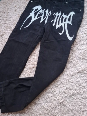 Revenge black denim jeans - Säljer ett par svarta jeans från Revenge med stort vitt tryck över framsidan. Jeansen har en rak passform och klassisk femficksdesign. Materialet är bomull och de har en edgy streetwear vibe.