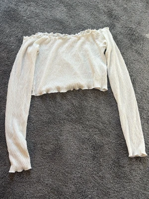 Vit offshoulder topp från BikBok - Säljer en söt vit offshoulder topp från BikBok i storlek M. Toppen är croppad med långa ärmar och har en ribbad struktur samt vågig kant upptill och nedtill. Perfekt för dig som gillar trendiga och feminina plagg.