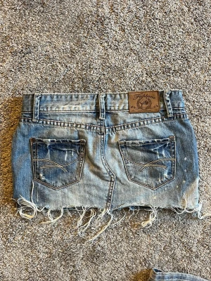 Blå jeanskjol med slitningar - Säljer en kort jeanskjol i blå denim med slitna detaljer och fransig nederkant. Kjolen har klassiska fickor fram och bak samt bälteshällor. Perfekt för dig som gillar en edgy och avslappnad stil. Saknar knapp men är super enkelt att fixa vilket jag även gjort förut! Storlek xs/s
