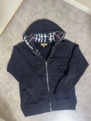 Svart hoodie från Burberry - Snygg svart hoodie från Burberry med dragkedja och klassiskt rutigt foder i huvan. Broderad logga på bröstet och två fickor framtill. Perfekt för dig som gillar exklusiv streetwear och vill ha något som sticker ut. Materialet känns mjukt och bekvämt.