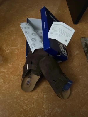 Bruna mockasandaler från Birkenstock - Säljer ett par bruna sandaler från Birkenstock i mjuk mocka. Modellen har en bred rem med metallknapp och öppen häl. Fotbädden är ergonomisk och sulan är i kork och gummi för extra komfort. Perfekta för dig som vill ha både stil och bekvämlighet.