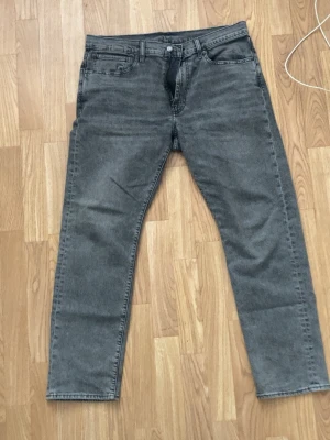 Grå jeans från Levi's 502 - Säljer ett par grå jeans från Levi's med klassisk femficksmodell och raka ben. Jeansen har en diskret tvättad look och är tillverkade i bomull med en liten läderpatch bak i midjan. Perfekta för en avslappnad och stilren outfit. Har en fläck vid ankeln efter misslyckad överföring av kastrull med mat från spis till matbord. Har inte tvättat de i tvättmaskinen. Köpta för 2 månader sedan. Inte använt speciellt mycket och vill därför bara bli av med de och säljer därmed för ett väldigt billigt pris.