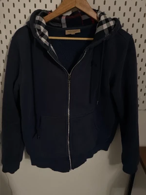 Mörkblå hoodie från Burberry - Snygg mörkblå hoodie från Burberry med dragkedja framtill och klassiskt rutigt foder i huvan. Hoodien har fickor på magen och ribbade muddar vid ärmslut och nederkant. Perfekt för dig som gillar stilrena och exklusiva plagg.