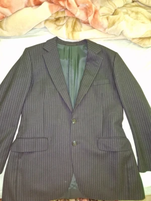 Mörkgrå randig kavaj med två knappar - Snygg mörkgrå kavaj med tunna vita kritstrecksränder och två knappar framtill. Klassisk passform med två fickor med lock och en bröstficka. Fodrad insida och smala slag ger en stilren look. Perfekt för dig som vill ha en tidlös och trendig kavaj.