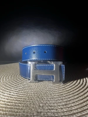 Blått skärp  - Snyggt blått bälte med ett stort, silverfärgatspänne. Bältet har vit kontrastsöm längs kanterna och är gjort i skinn. Perfekt accessoar för att lyfta din outfit.