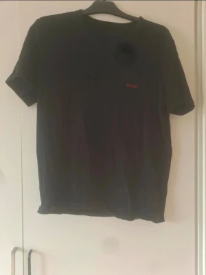 Svart t-shirt från BOSS - Säljer en svart t-shirt från BOSS med klassisk passform och korta ärmar. T-shirten har en enkel design med BOSS-loggan broderad i rött på bröstet. Perfekt för dig som gillar stilrena plagg med diskret branding.