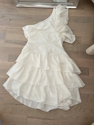 Vit asymmetrisk klänning med volanger - Säljer en vit klänning med ett axelband och volanger i flera lager. Klänningen har en asymmetrisk design med rynkad detalj på axeln och är tillverkad i ett lätt, strukturerat tyg. Perfekt för dig som gillar en unik och trendig look. Aldrig använd då den var för stor