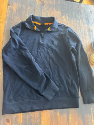 Mörkblå halfzip tröja från BOSS - Snygg mörkblå tröja från BOSS med halv dragkedja och hög krage. Tröjan har en diskret BOSS-logga på bröstet och orange detalj på insidan av kragen. Perfekt för en stilren och avslappnad look.