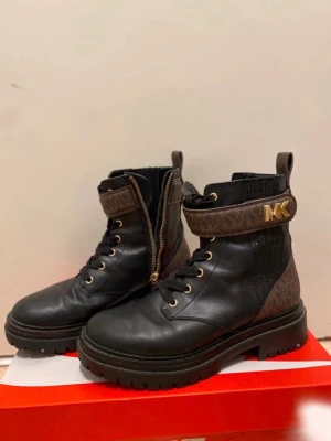 Svarta boots från Michael Kors - Snygga svarta boots från Michael Kors med bruna detaljer och guldfärgade MK-loggor på remmen. Boots i läder och textil med grov sula, snörning framtill och dragkedja på sidan. 