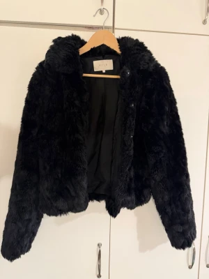 Svart pälsjacka från VILA - Snygg svart pälsjacka från VILA med mjuk fuskpäls och klassisk krage. Jackan har en rak, något kortare passform och stängs med dolda knappar framtill. Perfekt för att hålla värmen under kalla dagar och ge en trendig look.