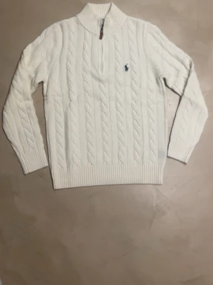 Vit kabelstickad tröja Polo Ralph Lauren - Kabelstickad vit tröja från Polo Ralph Lauren med halv dragkedja och hög krage. Tröjan har det klassiska blå broderade logotypen på bröstet och ribbstickade muddar vid ärmslut och nederkant. Perfekt för en stilren och avslappnad look.