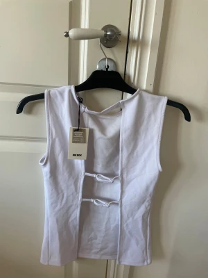 Vit ärmlös topp med knyt från Bik Bok - Snygg vit ärmlös topp från Bik Bok med två dekorativa knyt framtill. Toppen har en rak passform och är tillverkad i mjukt bomullsmaterial. Perfekt att styla med jeans eller kjol för en trendig look.