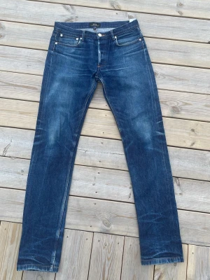 Blå jeans från A.P.C. - Säljer ett par klassiska blå jeans från A.P.C. i rak modell. Jeansen har fem fickor, silverfärgade nitar och knappgylf. Materialet är kraftig denim i 100% bomull med snygg tvätt och subtila slitningar. Perfekta för en stilren och avslappnad look.