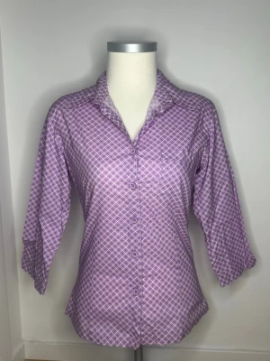 Lila rutig skjorta med trekvartsärm - Snygg lila skjorta med vitt rutmönster och trekvartsärm. Skjortan har klassisk krage, knappar framtill och en figurnära passform. Perfekt för dig som vill ha en färgglad och unik skjorta i garderoben. Passar även XS/S