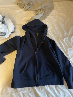 Mörkblå hoodie från Polo Ralph Lauren - Mörkblå hoodie med dragkedja från Polo Ralph Lauren. Klassisk design med huva, kängurufickor och röd broderad logga på bröstet. Mjuk insida och ribbade muddar. Perfekt för en avslappnad och stilren look.