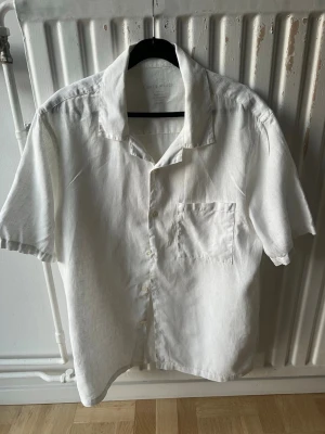 A Days March linneskjorta - Stilren vit skjorta i linne med korta ärmar och klassisk krage. Skjortan har en bröstficka och knäppning framtill. Perfekt för en avslappnad och fräsch look.