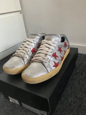 Silvriga sneakers med röda stjärnor - Unika sneakers i metallic silver med röda stjärnor på sidorna och blå detalj vid hälen. Skorna har vita snören och beige sula. Perfekt för dig som vill sticka ut med en cool och färgstark look. Storlek 39 passar 40. Hör av dig frågor och funderinga, byten kan vara intressant. Defekt på bild 6