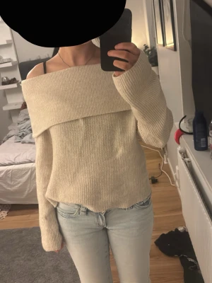 Beige off shoulder stickad tröja - Säljer en beige stickad tröja med off shoulder-design och bred krage. Tröjan har långa ärmar och är ribbstickad för en snygg och avslappnad look. Perfekt att styla med jeans eller kjol för en trendig outfit.