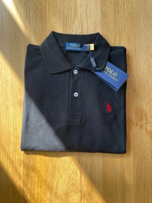 Svart polotopp från Polo Ralph Lauren - Säljer en klassisk svart polotopp från Polo Ralph Lauren med krage och knappar framtill. Toppens ikoniska röda logobrodyr på bröstet ger en snygg kontrast. Materialet är bomull och modellen har en normal passning.