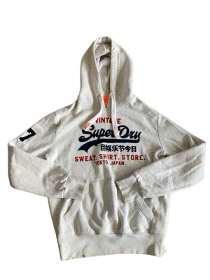 Grå hoodie från Superdry - Säljer en grå hoodie från Superdry med stor logga och japanska tecken på bröstet. Hoodien har huva med dragsko, ficka framtill och tryck i navy och rött..