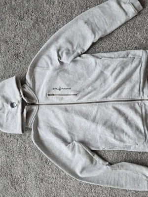 Ljusgrå hoodie från Sail Racing - Snygg ljusgrå hoodie från Sail Racing med dragkedja framtill och två sidofickor. Hoodien har en broderad logga på bröstet och en knappdetalj på huvan. Tillverkad i mjukt bomullsmaterial som känns skönt mot huden.