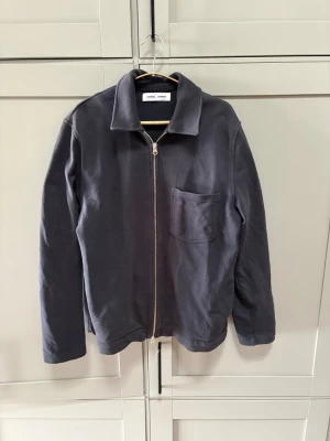Mörkblå overshirt från Samsoe Samsoe - Stilren mörkblå overshirt från Samsoe Samsoe med dragkedja framtill och en bröstficka. Overshirten har klassisk krage och är tillverkad i ett mjukt bomullstyg. Perfekt att slänga på sig över en t-shirt för en avslappnad look, Storlek M.
