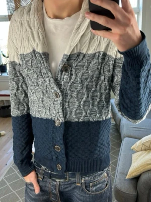 Flerfärgad kabelstickad cardigan strl M-L - Säljer en snygg kabelstickad kofta med breda blockränder i beige, grått och marinblått. Koftan har grovstickad struktur, sjalkrage och stängs med fem stora knappar framtill. Perfekt för lager på lager och chill stil. Väldigt unik och kommer från märket dreimaster. Storlek L men passar L-M. Hör av er vid frågor och funderingar!