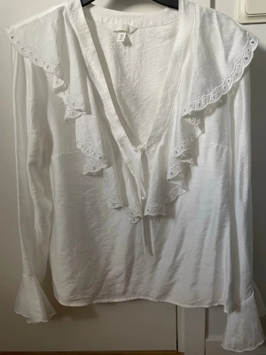 Vit blus med volang och spetsdetaljer - Vit blus från H&M med långa ärmar, djup v-ringning och volang med broderade spetsdetaljer längs ringningen och axlarna. Blusen har knytband framtill och lätt utsvängda ärmslut. Aldrig använd men utan prislapp