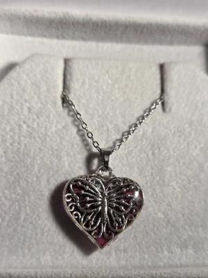 Halsband med hjärtformat hänge - Handgjort halsband med silverfärgad kedja i rostfritt stål och hjärtformat hänge. Hänget har en lila glittrig baksida och framsidan är dekorerad med en detaljerad silverfärgad fjäril och snirkliga mönster. Hänget lyser i mörker efter att ha varit i dagsljus eller solljus. Perfekt för dig som gillar unika accessoarer. Både hänget o kedjan tål vatten och missfärgas inte.