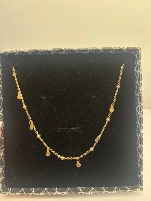Guldfärgat halsband med små hängen - Ett elegant guldfärgat halsband med flera små runda hängen och pärlliknande detaljer. Kedjan är tunn och smidig, perfekt för att ge en subtil men snygg touch till din stil. Levereras i en dekorativ ask.