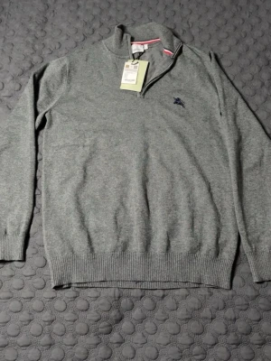 Grå halvzip tröja från Burberry - Snygg grå stickad tröja från Burberry med halvzip och broderad logga på bröstet. Tröjan har ribbade muddar och krage med diskret randig detalj på insidan. Perfekt för dig som gillar klassisk men ändå sportig stil. STORLEK M, PASSAR S. HELT NY PRISLAPP PÅ!!