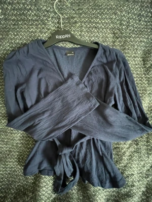 Mörkblå omlottblus Gina Tricot - Snygg mörkblå omlottblus från Gina Tricot med långa ärmar och knytband i midjan. Blusen har en lätt skrynklig struktur och djup V-ringning. Perfekt att styla med jeans eller kjol för en trendig look.