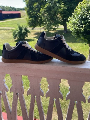 Maison margiela gats MMG - Stilrena svarta sneakers med vita skosnören och klassisk brun gummisula. Skorna har lågt skaft och ovandel i läder och mocka för en snygg kontrast. Perfekta för dig som gillar en tidlös och clean look. Pris kan alltid diskuteras😎😇