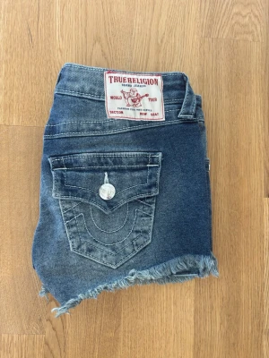 Blå jeansshorts från True Religion - Säljer ett par blå jeansshorts från True Religion med fransig kant och klassiska fickor. Shortsen har normal passform, låg midja och snygga slitningar framtill. Perfekta för sommaren och ger en avslappnad look.