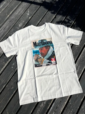 Beige / krämVit t-shirt med Marlboro-tryck vintage  - Beige /krämvit t-shirt med stort retroinspirerat Marlboro-tryck på bröstet. Motivet föreställer en cowboy i färg och texten 'Marlboro' upptill. Klassisk passform och rund halsringning. Perfekt för dig som gillar vintage vibes.