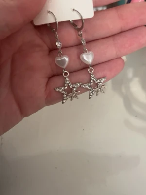 Silverfärgade örhängen med stjärnor - Säljer ett par snygga silverfärgade örhängen med hängande stjärnor dekorerade med små glittrande stenar och vita pärlhjärtan ovanför. Perfekta för dig som gillar unika detaljer och vill sticka ut lite extra.