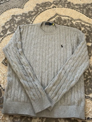 Grå kabelstickad tröja Polo Ralph Lauren - Snygg grå kabelstickad tröja från Polo Ralph Lauren med rund halsringning och klassisk broderad logga på bröstet. Tröjan har ribbade muddar vid ärmslut och nederkant och är tillverkad i mjukt material. Perfekt för en stilren look.