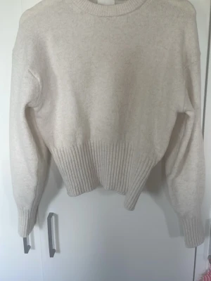 Beige stickad tröja med ribbad midja - Mysig beige stickad tröja med rund hals och bred ribbad mudd i midjan och vid ärmsluten. Tröjan har en avslappnad passform och långa ärmar. Perfekt för lager på lager och enkel att matcha med olika stilar.