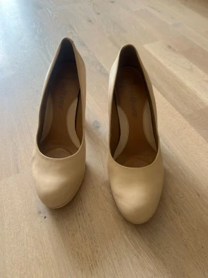 Beige klassiska pumps med platå-klack - Snygga beige pumps med rund tå och hög platå-klack. Skorna har en stilren design och är tillverkade i ett slätt material som ger en elegant look. Perfekta för dig som vill ha ett par neutrala klackar som passar till många outfits.