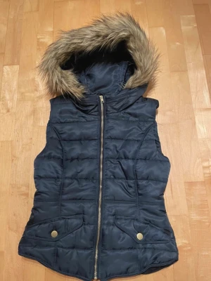Mörkblå dunväst med fuskpäls från H&M - Snygg mörkblå dunväst från H&M Divided med avtagbar huva och fuskpälsdetalj. Västen har dragkedja framtill, två fickor med tryckknappar och quiltad design. Perfekt för lager på lager och höst- eller vinteroutfits.